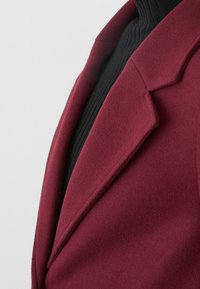 Burgunderfarbener Wollmischblazer mit Reverskragen, getragen über einem enganliegenden schwarzen Ripp-Turtleneck. Glatte Textur und maßgeschneiderte Verarbeitung.