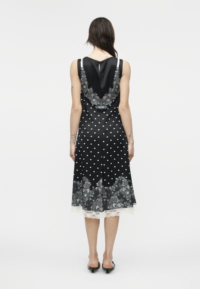 Γυναίκα που φορά μαύρο midi φόρεμα χωρίς μανίκια με λευκές πουά, λεπτομέρειες με δαντέλα λουλουδιών και μαύρα παπούτσια με χαμηλό τακούνι, από πίσω.