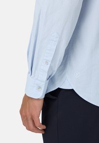 Camicia a righe di colore azzurro chiaro con maniche lunghe. Presenta un polsino con bottone e una parte posteriore sagomata, realizzata in un tessuto liscio.