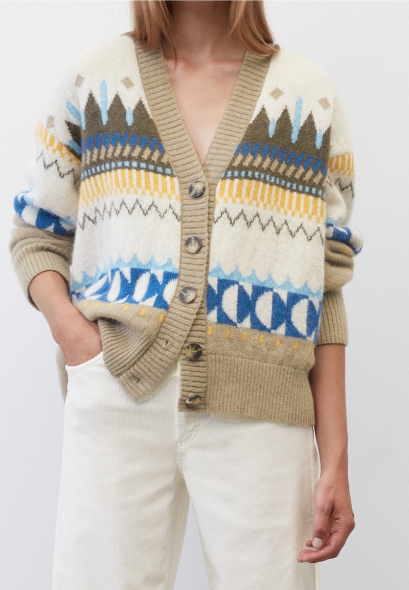 Femme portant un cardigan beige avec des motifs géométriques bleus, jaunes et olive, un pantalon crème, avec une main dans la poche sur un fond uni.