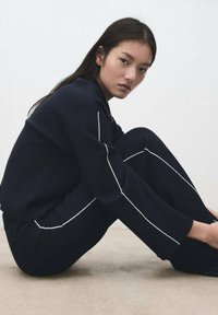 Tmavě modrý svetr s kalhotami, zdobený bílým lemováním podél švů. Měkká látka, volný střih. Model sedí na texturovaném povrchu.