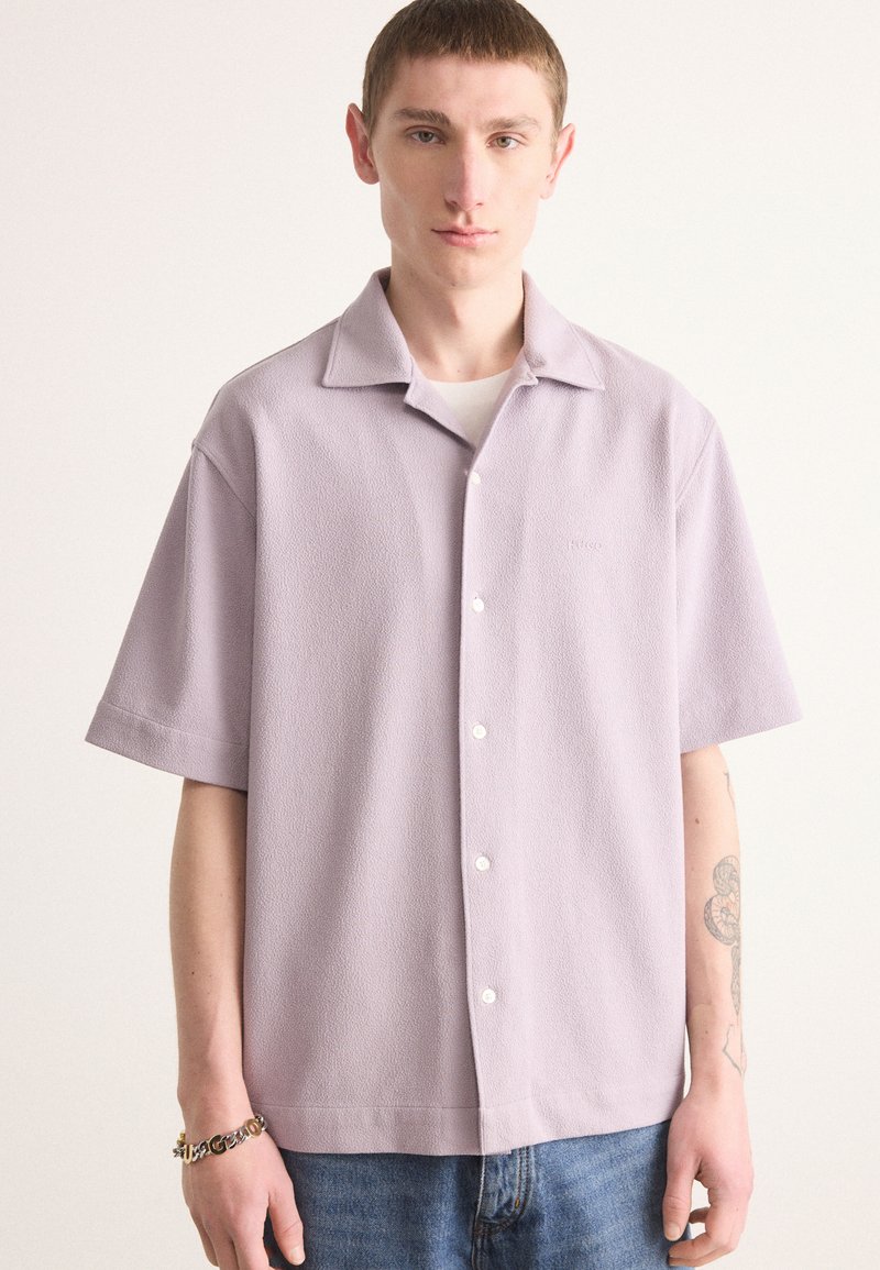 Kortärmad, ljus lavendelfärgad skjorta med knäppning, tillverkad av textil med struktur, med en avslappnad krage och en diskret logotyp på bröstet.