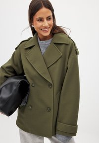 Olīvzaļš divrindu mētelis ar lieliem apakšlapiem, pogām un plecu akcentiem, apvienots ar pelēku džemperi un turēt melnu somu.