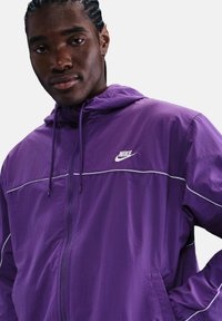 Giacca Nike viola con cappuccio, zip frontale, dettagli bianchi e tasche laterali; realizzata in materiale leggero e morbido.
