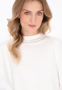 Witte gebreide trui met een hoge, geribbelde kraag. Soepele textuur, lange mouwen en een relaxed fit. Model heeft golvend blond haar en subtiele make-up.