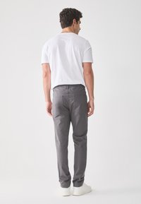 Un hombre de espaldas viste una camiseta blanca, pantalones grises y zapatillas blancas, de pie contra un fondo liso de color gris claro.
