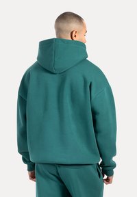 Sudadera de gran tamaño en color azul aguamarina, hecha de un tejido suave, con una gran capucha, puños acanalados y hombros caídos, vista desde la parte de atrás.