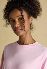 Light pink sweatshirt with a round neckline, soft texture, and simple design. Curly black hair partially frames the face. Neutral background.