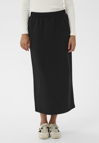 Personne portant une longue jupe noire avec une taille �élastique, un haut à longues manches blanc et des baskets blanches avec des rayures noires, se tenant sur un fond blanc.