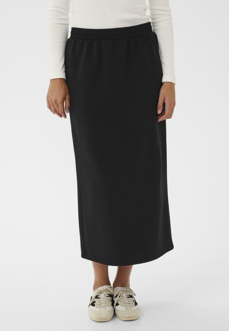 Personne portant une longue jupe noire avec une taille élastique, un haut à longues manches blanc et des baskets blanches avec des rayures noires, se tenant sur un fond blanc.