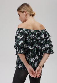 Off-shoulder svart bluse med blomstertrykk, volangermarmer og avslappet passform. Har grønne og hvite blomster mønstre mot en mørk bakgrunn.