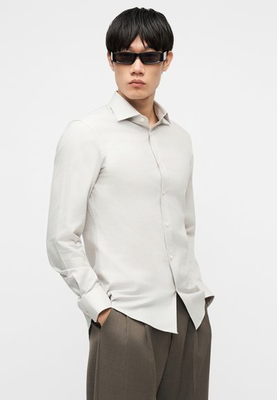 Mann iført hvit skjorte med knapper, taupe bukser og svarte, rektangulære solbriller, stående med hendene i lommene mot en hvit bakgrunn.