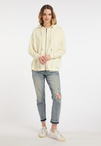 Hanorac crem cu fermoar și glugă, având șnururi și o croială lejeră. Asortat cu blugi deschiși la culoare, cu rupturi, și adidași albi.