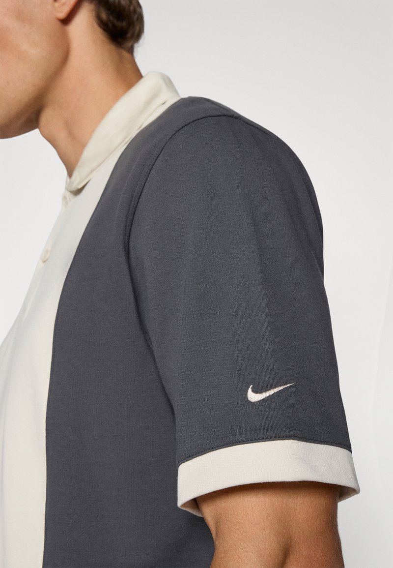 Camisa polo de cuello en gris oscuro y crema. Mangas cortas con puños en contraste. Presenta un pequeño logo de Nike en blanco en la manga izquierda.