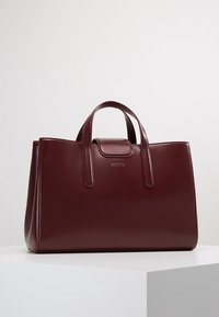 Bordeauxrode leren shopper met glanzende afwerking, rechthoekige vorm, twee bovenhandvatten, magnetische sluiting en geperst logo aan de voorkant.