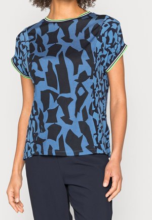 Woman wearing blue short-sleeve shirt with black abstract shapes and green-striped trim on collar and sleeve edges.