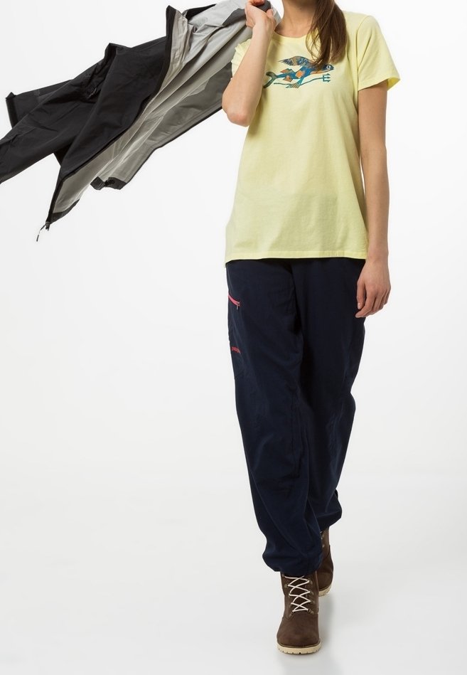 Žluté bavlněné tričko s modro-červeným grafickým motivem, doplněné o volné tmavě modré kalhoty a hnědé šněrovací boty. V ruce drží šedé a černé bundy.
