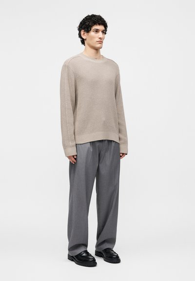 Homme aux cheveux bouclés foncés portant un pull beige texturé, un pantalon gris ample et des chaussures noires, debout devant un fond clair uni.