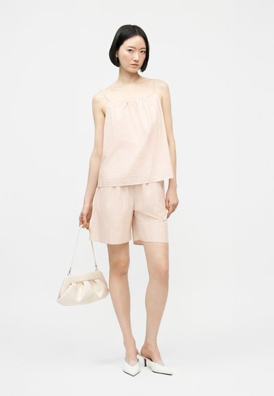 Žena s krátkymi čiernymi vlasmi má na sebe pásikavý púdrovo ružový top na ramienka a k nemu ladia krátke nohavice, drží v ruke bielohnedú kabelku a má na nohách biele lodičky.