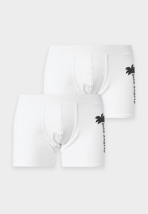 Junger Mann trägt weiße Boxershorts mit schwarzem Palmenmotiv, steht barfuß vor einem schlichten, hellen Hintergrund.