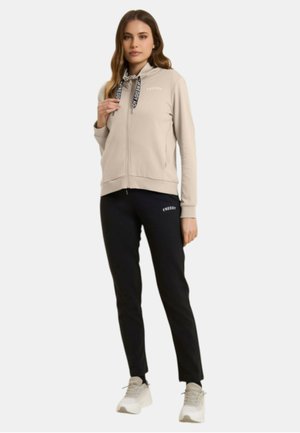 Donna che indossa una felpa con zip beige con coulisse nere e pantaloni aderenti neri, in piedi, con sneakers beige su sfondo bianco.