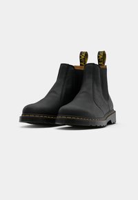 Bottes Chelsea en cuir noir avec surface texturée, panneaux latéraux élastiques et languettes de traction jaunes. Elles présentent une semelle en caoutchouc épaisse avec des surpiqûres jaunes.