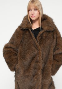 Brūna mākslīgā kažokāda ar plašu apkakli, greznu tekstūru un brīvu piegriezumu. Iespējas ietver lielas piedurknes un vienu aizdari priekšpusē.