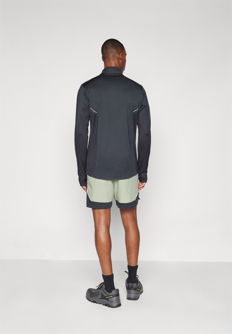 Top negro de manga larga con acentos reflectantes, combinado con pantalones cortos verde claro y zapatos grises, visto desde la parte posterior.