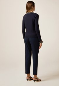 Vrouw in een marineblauwe top met lange mouwen en een gebloemde kraag, passende marineblauwe broek, zwarte armbanden en zwarte lage haksandalen, staand met haar rug naar de camera.