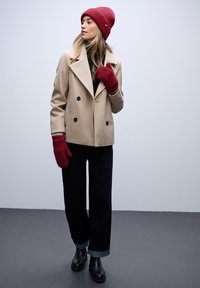 Beige dubbelzijdig gevaste jas, zwarte coltrui, donkere jeans met opgerolde manchetten, rode gebreide beanie en handschoenen, zwarte enkellaarsjes. Gladde texturen.