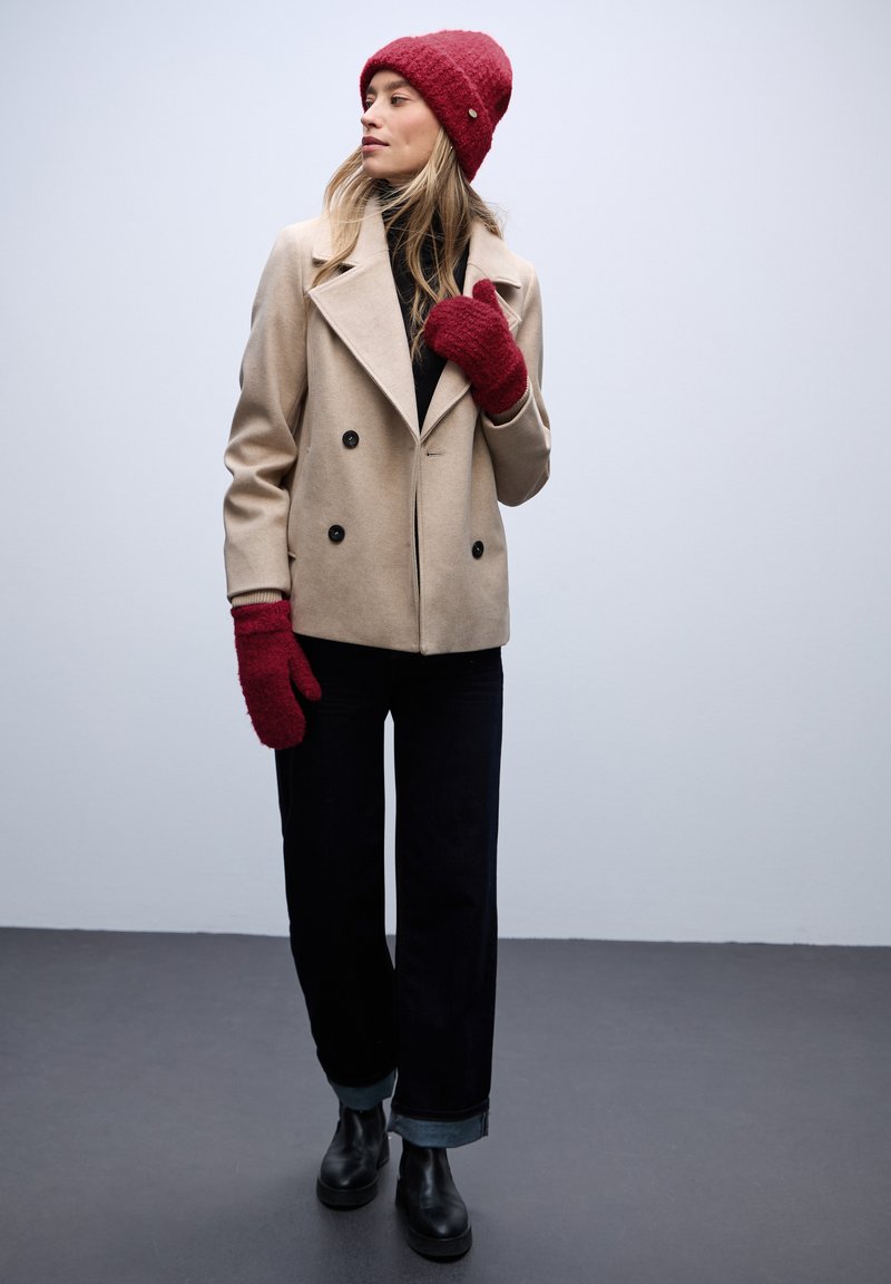 Beige dubbelzijdig gevaste jas, zwarte coltrui, donkere jeans met opgerolde manchetten, rode gebreide beanie en handschoenen, zwarte enkellaarsjes. Gladde texturen.