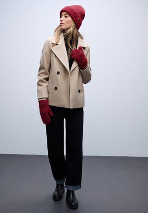 Beige double-breasted coat, black turtleneck, dark jeans with rolled cuffs, red knit beanie and gloves, black ankle boots. Smooth textures.