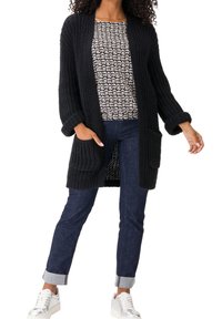 Schwarze, gestrickte Strickjacke mit offenem Front und zwei Taschen, kombiniert mit einem gemusterten Oberteil und dunklen Jeans. Das Model trägt weiße Sneaker.