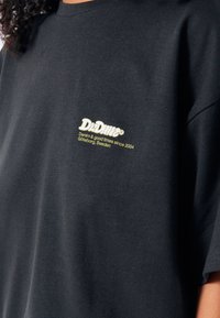 Černé bavlněné tričko s žlutou vyšitou nášivkou na levém prsu, text zahrnuje "Dr Denim" a údaje o lokaci. Krátké rukávy, volný střih.