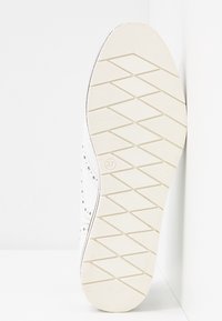 Semelle d'une chaussure blanche avec des rainures diagonales pour la traction, indiquant la taille 37 à l'intérieur d'un cercle près du centre.