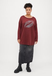 Top de mangas longas cor bordô com uma estampa de lábios prateados, combinado com uma saia de ganga preta e botas pretas de sola robusta, em fundo branco liso.