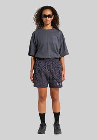 Sivá oversized tričko, vzorované šortky s geometrickými vzormi, čierne športové topánky a tmavé slnečné okuliare. Minimalistický štýl.