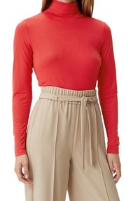 Vrouw in een rode lange mouw turtleneck, gestoken in hooggetailleerde beige broek met een stoffen riem die in een knoop om de taille is gebonden.