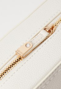 Weiße, strukturierte Leder-Tasche mit einem goldenen Reißverschluss, verziertem Beschlag und einem klaren Kristallakzent am Reißverschluss-Anhänger.