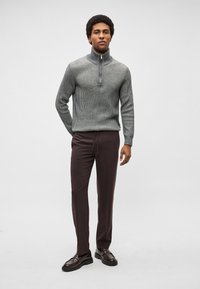 Pulôver cinzento canelado com gola meia-canelada, combinado com calças cónicas castanho-escuras e mocassins pretos lisos. Modelo em pé contra um fundo neutro.