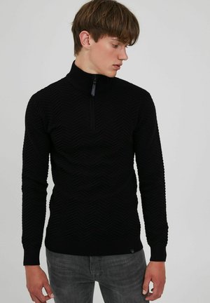 Schwarzer Strickpullover mit hohem Kragen und teilweisem Reißverschluss. Besitzt ein strukturiertes Zickzackmuster und gerippte Bündchen.