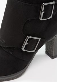 Bottine en daim noir avec deux boucles argentées et un talon bloc texturé.