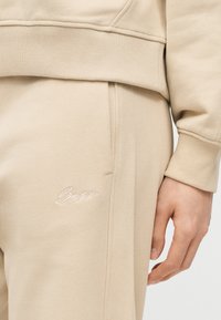 Μπεζ σετ φούτερ και παντελόνι φόρμας με ριμπ μανσέτες και μέση, με κεντημένο λογότυπο "Boss" στο αριστερό μηρό, χέρι ακουμπισμένο στο πλάι.