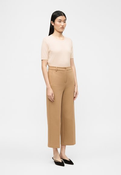 Γυναίκα που στέκεται φορώντας ένα ανοιχτό μπεζ τοπ με κοντά μανίκια, καμηλό παντελόνι με φαρδιά μπατζάκια μέχρι τους αστραγάλους και μαύρα φλατ παπούτσια με μύτη σε σχήμα μυτερού και λουρί στη φτέρνα.