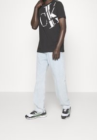 Svart t-shirt med vitt logotypdesign, ljusblåa löst sittande jeans och svarta, vita och gröna sneakers med mönstrade sulor.