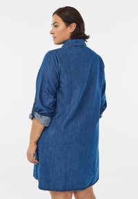 En kvinna med axellångt mörkt hår bär en denimklänning, sedd bakifrån, med uppkavlade ärmar och en avslappnad passform.
