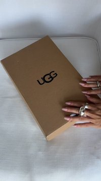 Hnědá kartonová krabice s černým logem "UGG" na vrchu. Ruce jsou položeny na krabici, která leží na světlém strukturovaném povrchu.