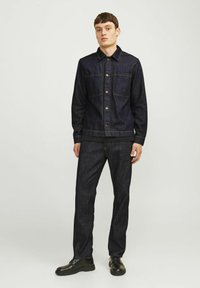 Chaqueta de mezclilla oscura con frente abotonado, dos bolsillos en el pecho y cuello. Combinada con pantalones de mezclilla oscuros y zapatos negros con cordones.