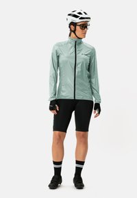 Lichtgroene, lange mouwen fiet jacket met een rits aan de voorkant, gecombineerd met zwarte shorts. Zwarte fietsschoenen en vingerloze handschoenen worden gedragen.