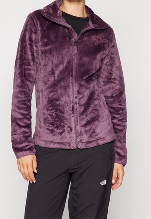 Personne portant une veste zippée violette en tissu pelucheux et un pantalon noir avec le logo The North Face sur les deux vêtements.